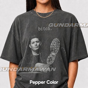 May include: A charcoal gray short-sleeve t-shirt in a pepper wash finish. The front features a black and white photographic print of a man with a boot sole graphic overlaid, accompanied by the lowercase text "bitch." printed in a simple serif font.