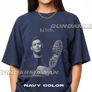 May include: A navy blue short-sleeve t-shirt featuring a graphic print of a man and a boot sole. The text "bitch." is printed above the man's head. The shirt has a crew neckline and a loose, boxy fit, showcasing a casual and simple design.