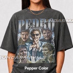 May include: A charcoal gray short-sleeve t-shirt featuring a graphic print of actor Pedro Pascal in various roles. The design includes the text "PEDRO Pascal" in a bold font, layered over lightning bolt imagery. The shirt has a crew neckline.