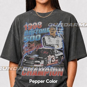 May include: A charcoal grey short-sleeve t-shirt featuring a graphic print of the 1998 Daytona 500, a portrait of Dale Earnhardt Jr., and his number 3 race car. The design includes the text 1998 Daytona 500, Dale Earnhardt Jr., and Champion.
