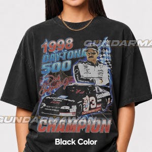 Peut inclure: Un t-shirt noir avec une impression graphique du Daytona 500 de 1998, Dale Earnhardt Jr. et sa voiture de course num&eacute;ro 3. Le design comprend des &eacute;clairs bleus, un drapeau &agrave; damier et le texte 1998 DAYTONA 500 et CHAMPION en rouge et bleu.