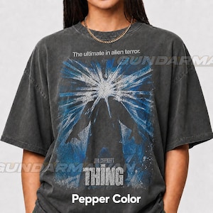 May include: A charcoal gray short-sleeve t-shirt featuring a graphic print from the movie The Thing. The design displays a blue and white starburst effect behind a dark silhouette with the text The ultimate in alien terror and John Carpenter's THE THING.