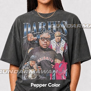 May include: A charcoal grey short-sleeve t-shirt featuring a graphic print with the text DARIUS Day. The design includes multiple photographic portraits of a person, one wearing glasses and a sweater, others in a plaid shirt or a red jacket.