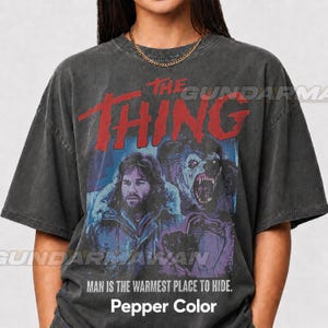 Pode incluir: Uma t-shirt cinzento-carvão de manga curta com uma impressão gráfica do filme The Thing. O design inclui o texto vermelho THE THING, um retrato de uma personagem barbuda, uma criatura e o texto MAN IS THE WARMEST PLACE TO HIDE.