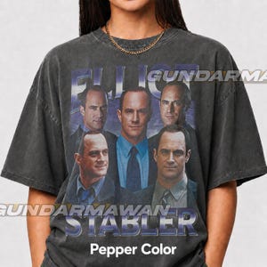 Pode incluir: Uma t-shirt cinzento-carvão com lavagem vintage, apresentando uma impressão gráfica do ator Christopher Meloni em várias poses. O texto ELLIOT STABLER está impresso numa fonte azulada e arrojada na parte superior e inferior.
