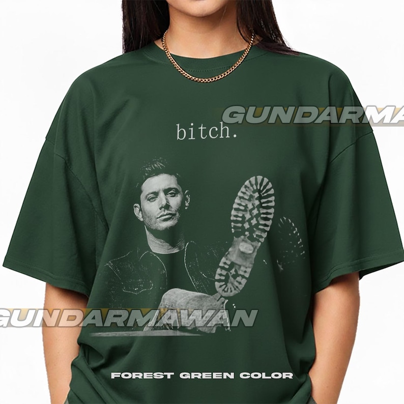 May include: A forest green short-sleeve t-shirt featuring a black and white graphic of a man and a boot sole. The text "bitch." is printed above the man's head. The shirt has a crew neckline and a loose, boxy fit with a solid dark green color.