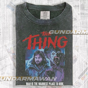 May include: Dark gray Comfort Colors t-shirt featuring a graphic from the movie "The Thing." The design includes the title in red, and an image of a man and a creature. The text "MAN IS THE WARMEST PLACE TO HIDE" is at the bottom.