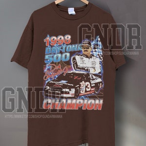 Peut inclure: Un t-shirt marron &agrave; manches courtes avec une impression graphique du Daytona 500 de 1998, Dale Earnhardt Jr. et une voiture de course noire portant le num&eacute;ro 3. Le texte indique 1998 DAYTONA 500, Dale Earnhardt Jr. et CHAMPION en lettres grasses.