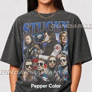 May include: A charcoal gray short-sleeve t-shirt featuring a graphic print with the text STUCKY at the top. The design includes multiple images of two characters, one wearing a Captain America suit and the other with long hair, in various poses.