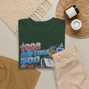 Peut inclure: Un t-shirt vert fonc&eacute; avec une impression graphique du Daytona 500 de 1998. Le motif comprend le texte 1998 DAYTONA 500 en rouge et bleu, ainsi qu'une illustration d'un pilote de course portant une casquette et des lunettes de soleil, avec un drapeau &agrave; damier.