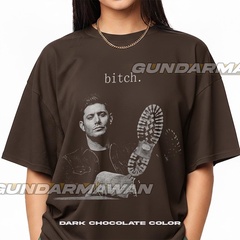 May include: A dark chocolate brown short-sleeve t-shirt featuring a graphic print of a man and a boot sole. The text "bitch." is printed above the man's head. The shirt has a crew neckline and a loose, boxy fit, showcasing a monochromatic design.