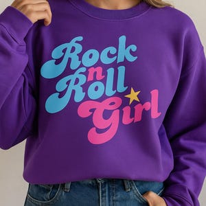 Puede incluir: Una sudadera morada con el texto "Rock n Roll Girl" en azul, rosa y amarillo. La sudadera es de color liso y tiene cuello redondo. El texto está en una fuente retro de estilo burbuja.