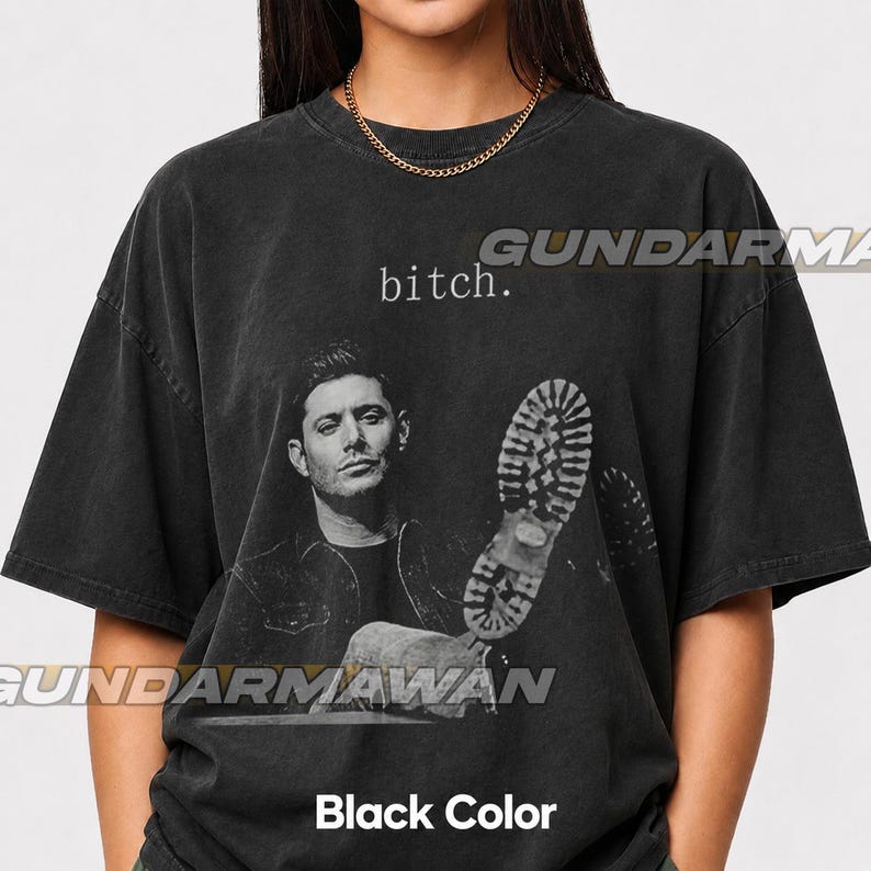 May include: A black short-sleeve t-shirt featuring a monochromatic graphic of a man and a large boot print. The text "bitch." is printed in white above the man's head. The shirt has a crew neckline and a faded, vintage-style wash finish.