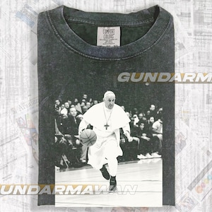 May include: Dark gray Comfort Colors t-shirt featuring a black and white image of the Pope dribbling a basketball. The shirt has the text "GUNDARM" and "UNDARMAWAN" printed on it. The shirt has a crew neck and short sleeves.