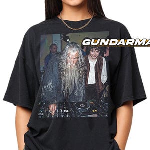 May include: Black t-shirt featuring a graphic of two characters from The Lord of the Rings behind a DJ mixing board. The text "GUNDARMA" is printed in white on the upper right side of the shirt.