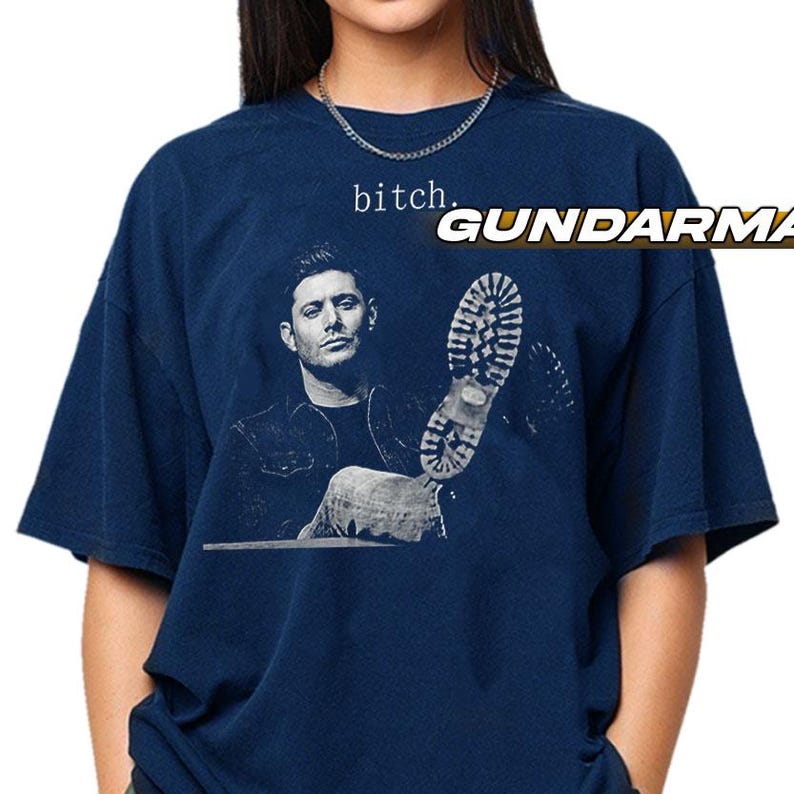 May include: A navy blue short-sleeve t-shirt featuring a monochromatic graphic of a man and a boot sole. The text "bitch." is printed above the man's head in a simple font. The shirt has a crew neckline and a loose, relaxed fit.