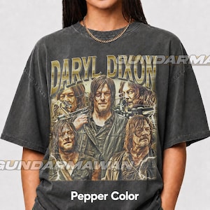 May include: A charcoal gray short-sleeve t-shirt featuring a graphic print of the character Daryl Dixon. The design includes the text DARYL DIXON in gold lettering above multiple images of the character holding a crossbow. The shirt is pepper color.