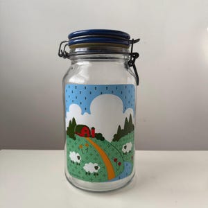May include: A clear glass jar with a blue lid and a metal clasp. The jar features a colorful farm scene with a red barn, green hills, white sheep, and a blue sky with clouds. The jar is designed for storage.