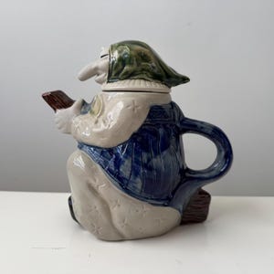 May include: A ceramic teapot shaped like a figure with a long nose, wearing a green headscarf, a blue vest, and a light-colored garment with star-shaped patterns. The figure holds a brown object, and the teapot features a curved handle and spout.