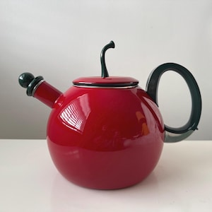 May include: A glossy red teapot with a rounded body, a dark green handle, and a matching spout. The lid has a dark green stem-like handle. The teapot is designed for brewing tea and has a modern, minimalist aesthetic.