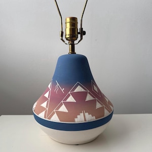 May include: A ceramic table lamp base with a bell-shaped silhouette, featuring a gradient color transition from blue at the top to a dusty pink and tan midsection, accented with white geometric line patterns and a solid white base section.