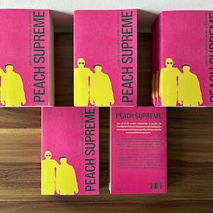 Op de afbeelding: Een stapel roze paperback boeken met de titel "Peach Supreme" en de namen van de auteurs Donovan Bridgeman en Nicholas Hiddison. De omslag toont een zwart en geel silhouet van twee personen.