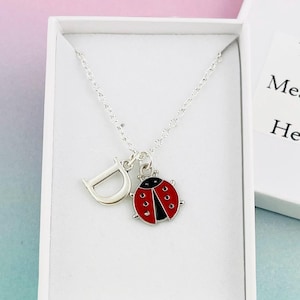 Puede incluir: Un collar de plata con un dije con la letra D y un dije de mariquita roja. El collar está en una caja blanca con el texto "Tu mensaje aquí".