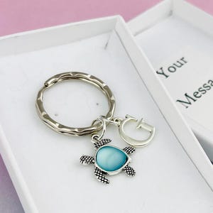 May include: A silver key ring with a blue turtle charm and a silver letter G charm. The keyring is in a white gift box with the text "Your Message Here".
