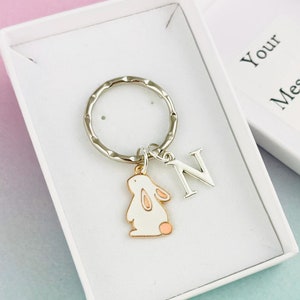 Puede incluir: Una caja de regalo blanca con un llavero plateado con un dije de conejo blanco y una letra N plateada. La caja tiene el texto "Your Message Here" impreso en la parte superior.