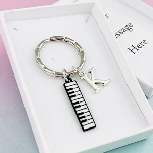 Könnte beinhalten: Ein silberner Schlüsselanhänger mit einem Klaviertasten-Anhänger und einem silbernen Buchstaben K-Anhänger. Der Schlüsselanhänger befindet sich in einer weißen Geschenkbox mit dem Text "Message Here".