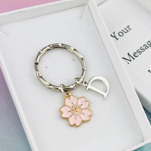 Puede incluir: Una caja de regalo blanca con un llavero plateado con un dije de flor rosa y una letra D plateada. La caja tiene el texto "Your Message Here" impreso en la tapa.