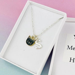Puede incluir: Un collar plateado con un colgante de gato negro y un charm con la letra "S" plateada, presentado en una caja de regalo blanca abierta. La caja tiene una pegatina que dice "Your Message Here" sobre un fondo degradado rosa y azul.