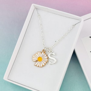 Peut inclure: Une boîte cadeau blanche contenant un collier en chaîne argent avec un pendentif en forme de marguerite et un pendentif en forme de lettre 'S'. La boîte a un espace vide pour un message personnalisé qui dit "Votre message ici".