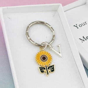 Peut inclure: Une boîte cadeau blanche contenant un porte-clés argenté avec un charme en forme de tournesol et la lettre "V". La boîte a un espace vide pour un message personnalisé.
