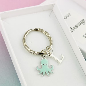 May include: A silver keyring with a light blue octopus charm and a silver letter L charm. The keyring is in a white gift box with the text "our message here" printed on the box.