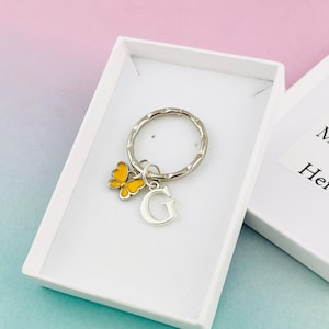 Op de afbeelding: Een zilveren sleutelhanger met een gele vlinderbedel en een zilveren letter G-bedel. De sleutelhanger zit in een wit cadeaudoosje met een wit kaartje waarop staat "Uw bericht hier".