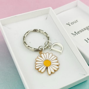 Op de afbeelding: Een witte cadeaudoos met een zilveren sleutelhanger met een witte madeliefje-bedel en een geel hart. De doos heeft de tekst "Uw bericht hier" op de bovenkant.