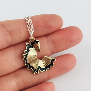 May include: A gold and black pencil sharpener charm necklace. The charm is shaped like a pencil point and has a black outline.