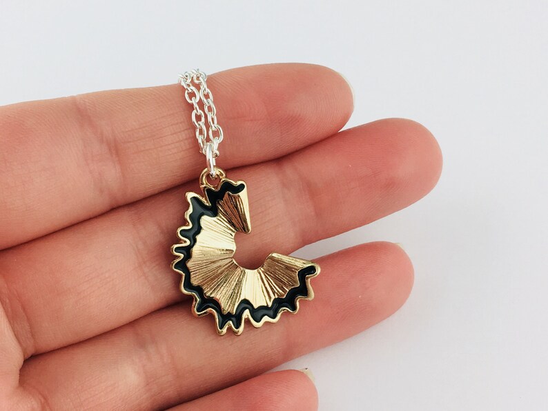 May include: A gold and black pencil sharpener charm necklace. The charm is shaped like a pencil point and has a black outline.