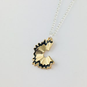 May include: A gold necklace with a black and gold pencil sharpener pendant. The pendant is shaped like a pencil sharpener with a black rim and gold shavings.