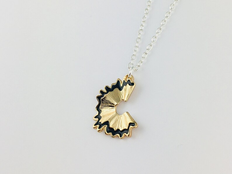 May include: A gold necklace with a black and gold pencil sharpener pendant. The pendant is shaped like a pencil sharpener with a black rim and gold shavings.