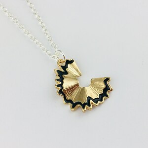 May include: A gold necklace with a black and gold pencil sharpener pendant. The pendant is shaped like a pencil sharpener with a black outline and a gold interior.