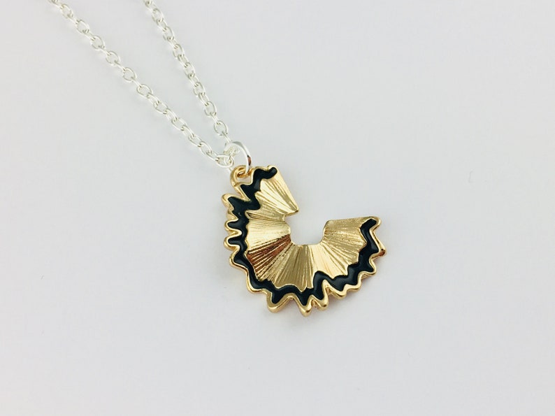 May include: A gold necklace with a black and gold pencil sharpener pendant. The pendant is shaped like a pencil sharpener with a black outline and a gold interior.