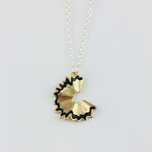 May include: A gold necklace with a pencil sharpener pendant. The pendant is black and gold and shaped like a pencil sharpener with a black edge.