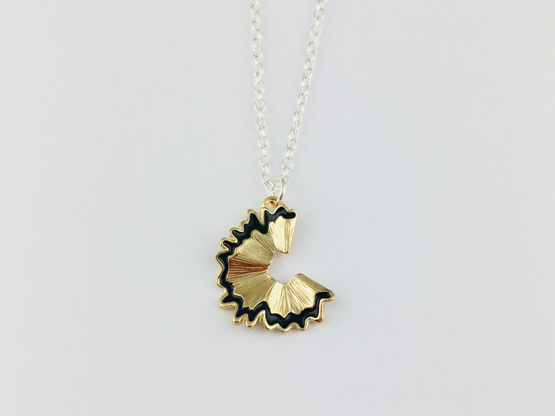 May include: A gold necklace with a pencil sharpener pendant. The pendant is black and gold and shaped like a pencil sharpener with a black edge.