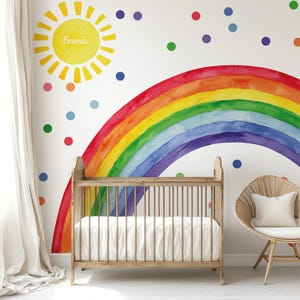 Könnte beinhalten: Eine Szene im Kinderzimmer mit einem Aquarell-Regenbogen, einer Sonne mit dem Namen "Emma" und bunten Punkten an einer weißen Wand. Ein Holzbettchen und ein Korbstuhl mit einem weißen Kissen vervollständigen die fröhliche Einrichtung.