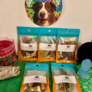 May include: Several simmer pot sachets in clear pouches with orange and blue detailing. The sachets contain dried herbs and spices. A round tie-dye sign with a dog's face and the text "Bear's Best Wishes" is above the sachets.