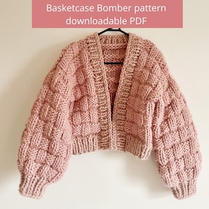 Puede incluir: Una chaqueta bomber de punto rosa con un patrón de cesta. La chaqueta tiene manga larga y un corte holgado.