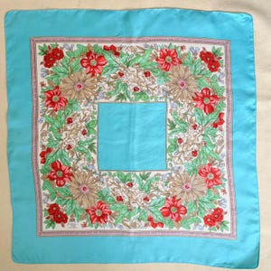 May include: A turquoise blue silk scarf with a floral pattern. The pattern features red, white, and green flowers with a light brown center. The scarf has a light pink border.
