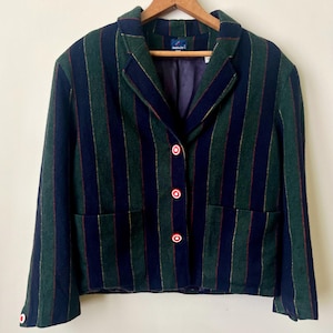 May include: A green and navy blue striped blazer with red-trimmed white buttons. The blazer has a classic collar, two front pockets, and is hanging on a wooden hanger. The lining is purple.